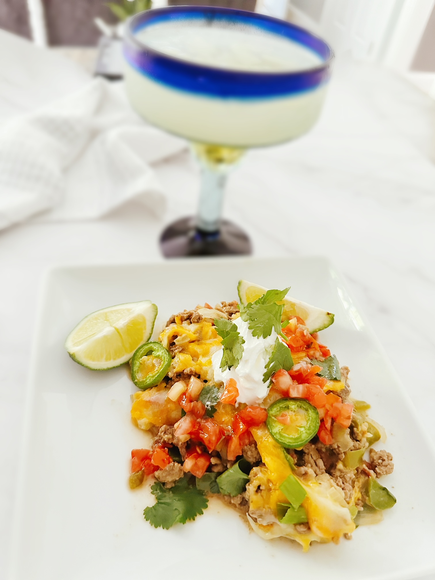 keto mexican taco casserole on a white dish with fresh lime, sour cream, and cilantro with a margarita in the background