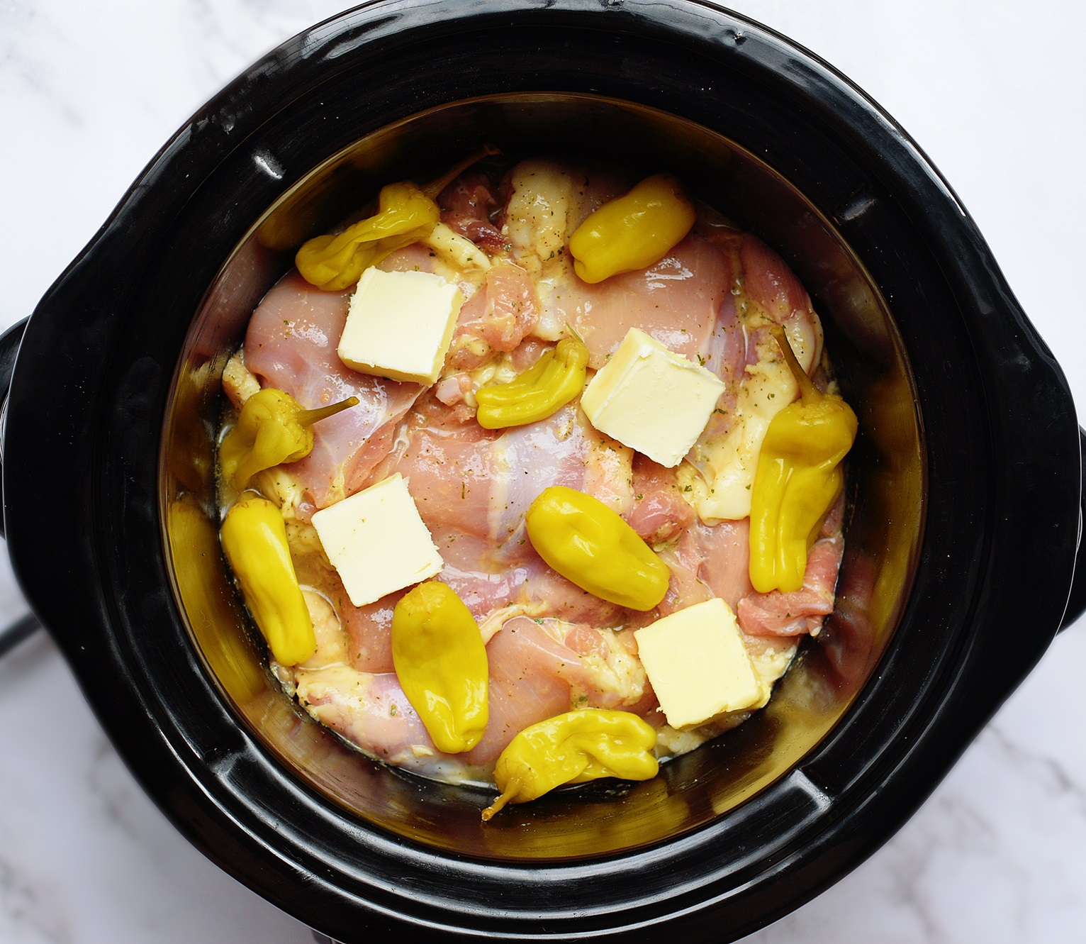 raw chicken topped with broth, ranch seasoning, pepperoncini and butter cooking in a crock pot