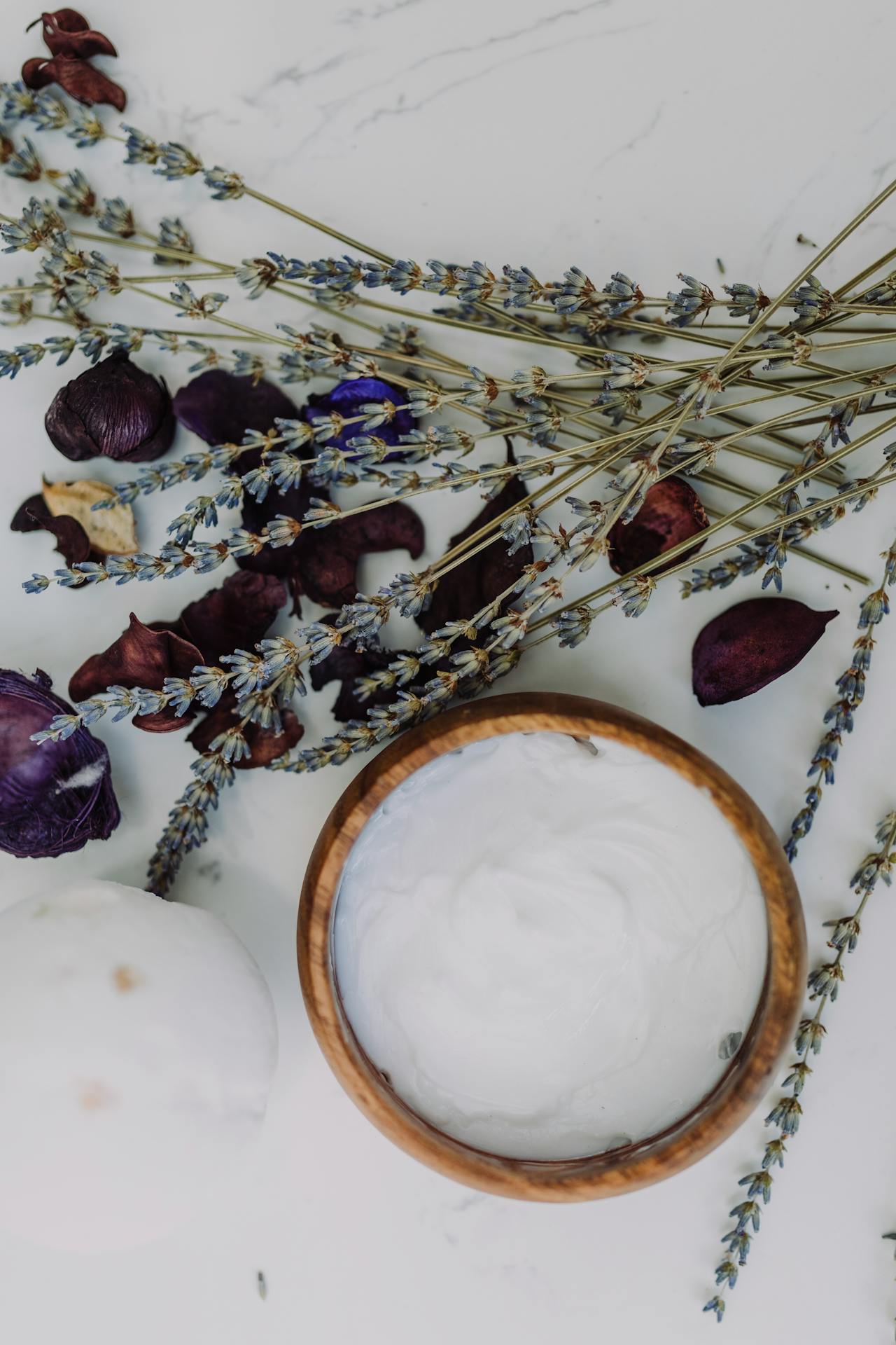 fresh herbs and petals next to white lotion in a wood bowl