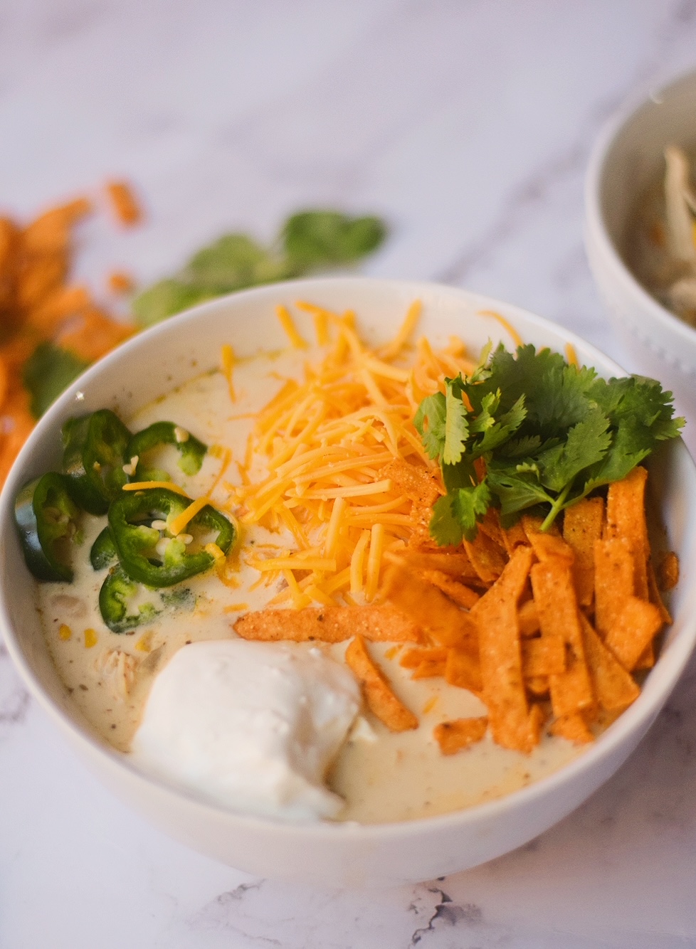 white chicken chili topped with cheese, cilantro, cream cheese, tortilla strips and jalapeno peppers in a white bowl