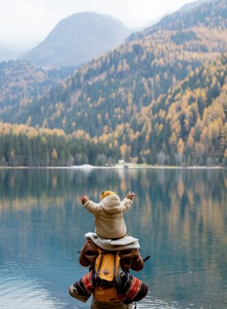 baby on dads shoulders in front of a lake with mountains in the background