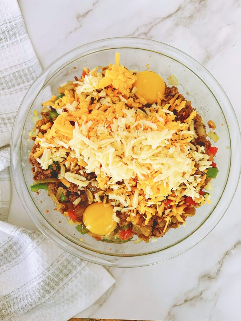 cooked ground beef, diced tomatoes, green bell pepper, onion, and garlic mixed together in a glass bowl topped with raw eggs and cheese ready to be mixed