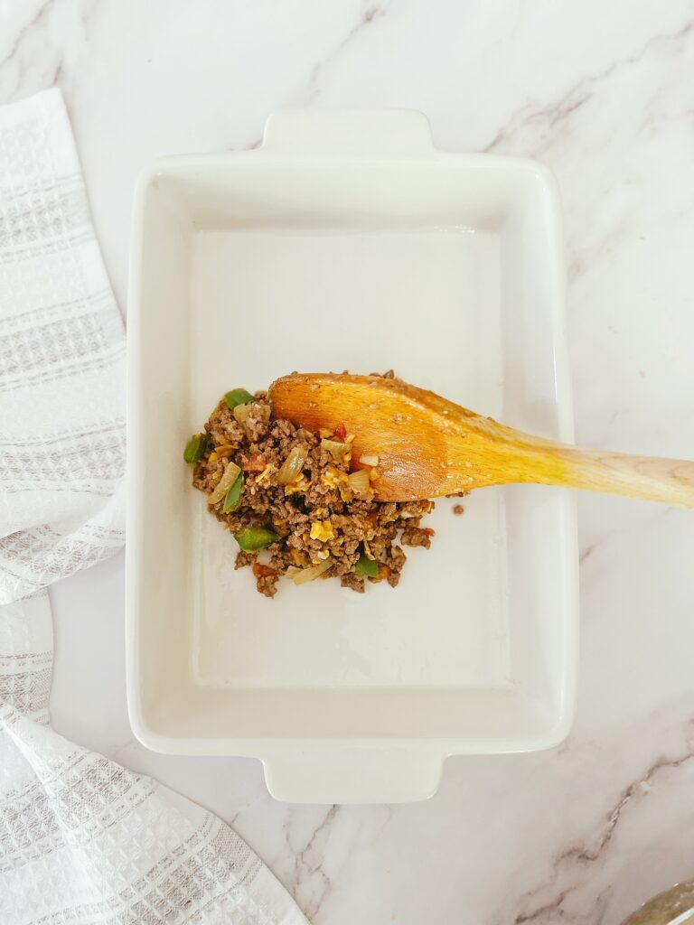 wooden spoon placing keto mexican casserole filling into a white porcelain baking dish