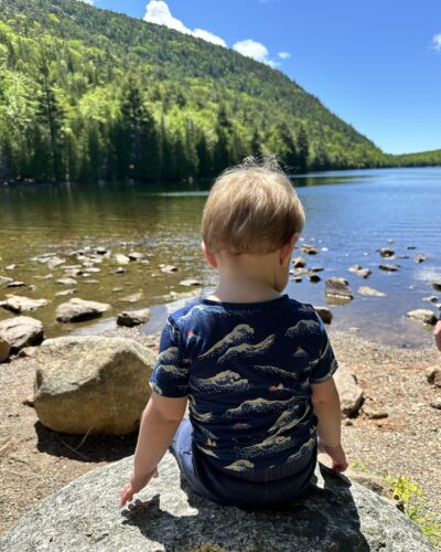 toddler facing a lake sitting on a rock